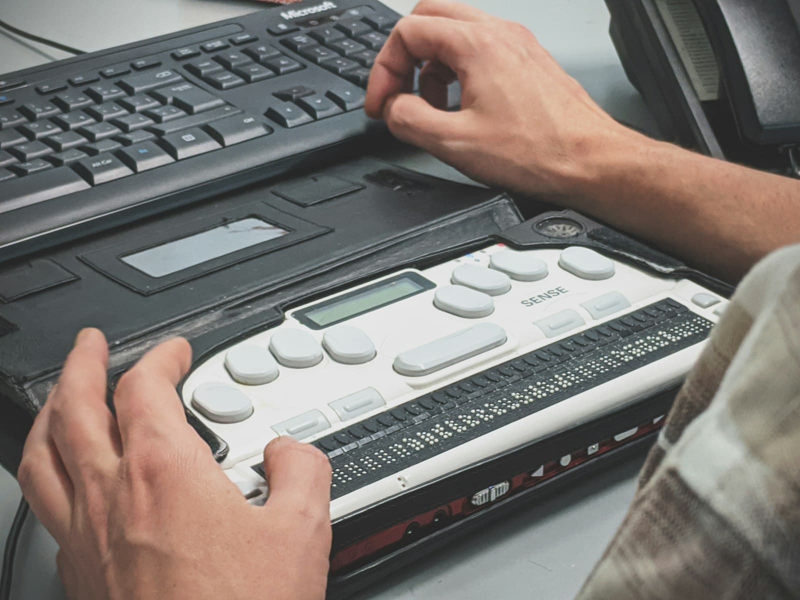 Close-up of hands on a refreshable braille display beside a laptop running a screen reader.